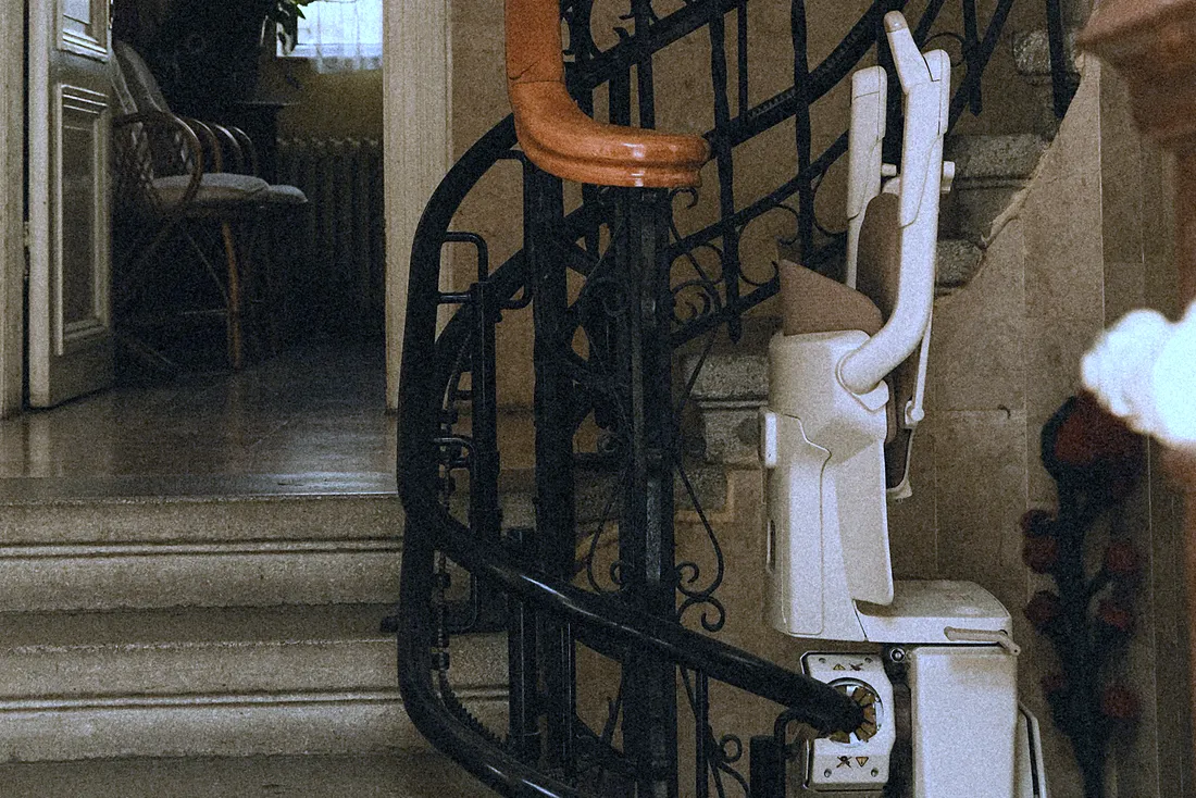 Siège de monte-escalier replié au pied d'un escalier tournant à rampe en fer forgé, dans le hall d'une maison ancienne