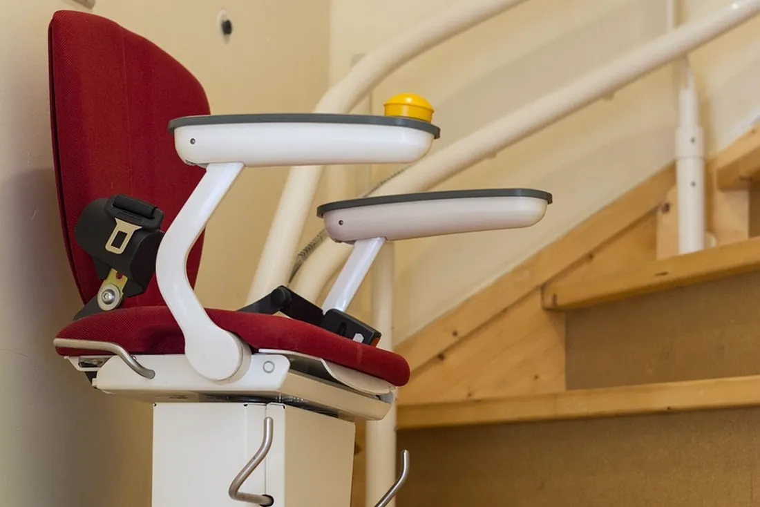 Fauteuil rouge de monte-escalier vu de côté, accoudoirs blancs, sur un rail blanc cintré qui suit un escalier tournant en bois